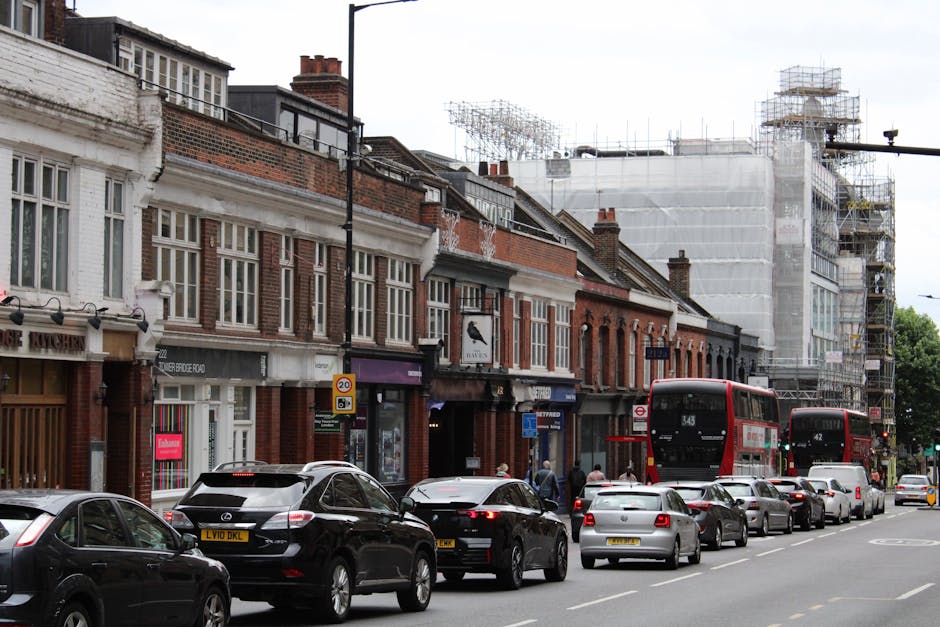A white moving van is parked on a city street in front of a large, multi-storey brick building with numerous windows and decorative architectural details. The van is positioned near the sidewalk, with
