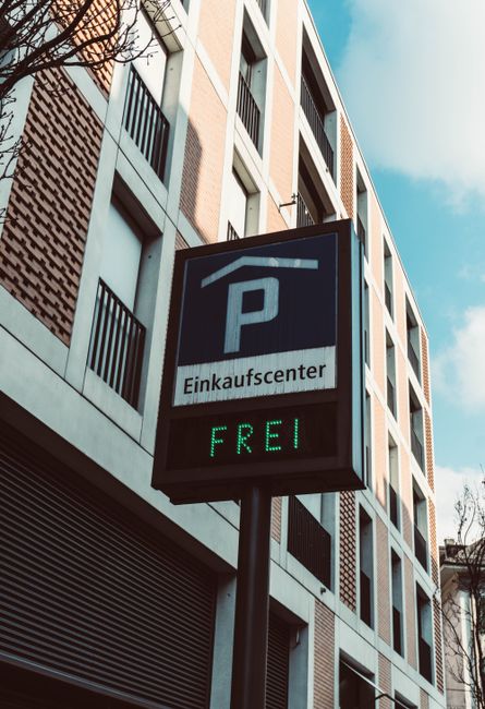 A vertical parking sign mounted on a pole in front of a modern multi-storey building with a facade featuring white and brick-colored panels and black window frames. The sign displays a large white 'P'
